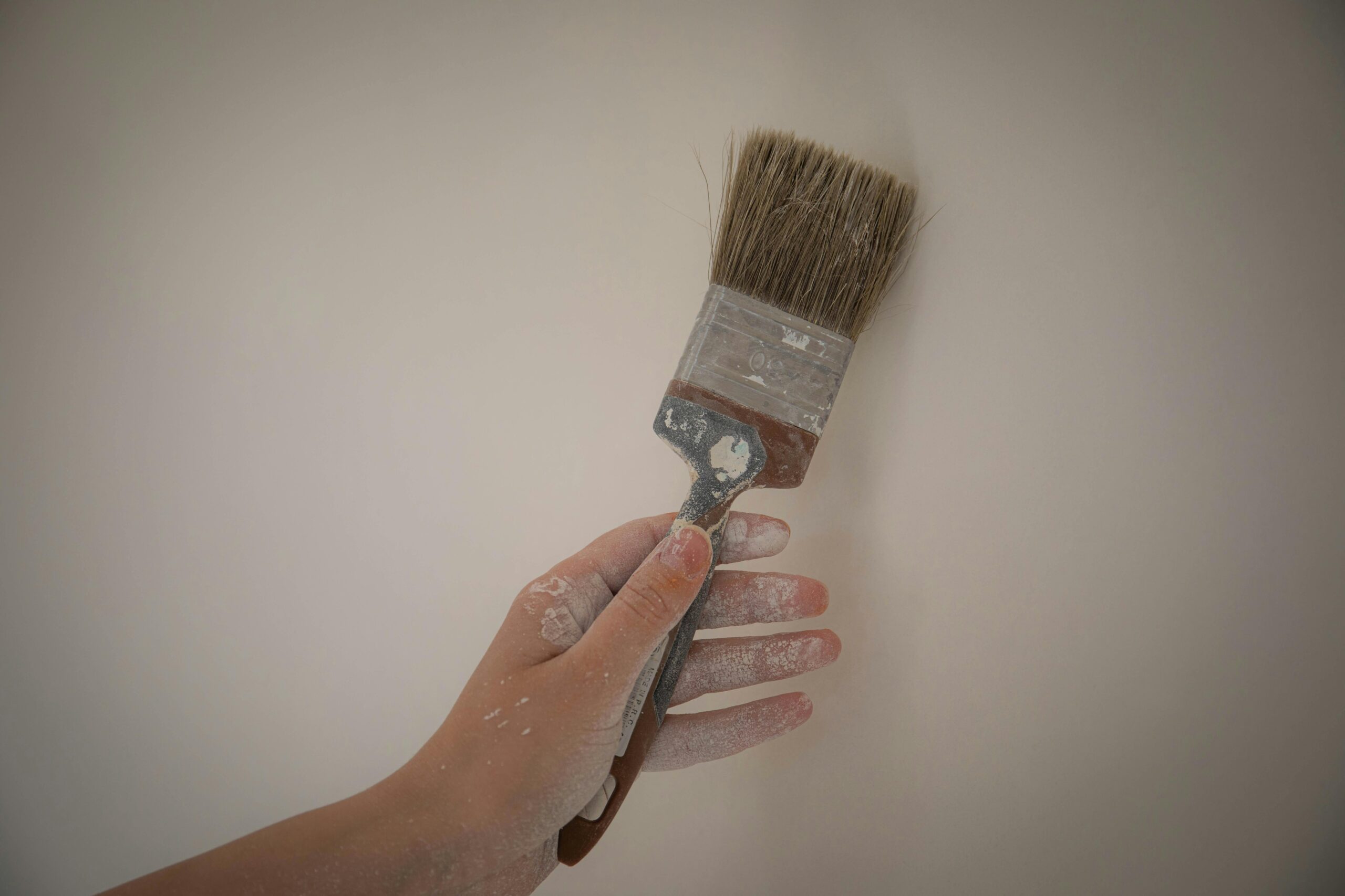 A hand holds a paintbrush with white paint against a plain wall, showcasing DIY home improvement.